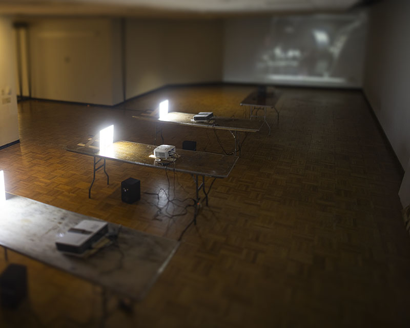 Temporary home screen image.

An image looking into a room, shot from its top-right corner with a tilt-shift lens. In the room there are four tables, spaced out. Three closer tables are perpendicular to the camera and each have a projector and small screen, which is reflecting enough light to light the space. The images are too bright to be captured by the camera. The fourth table is furthest away and pointed towards a far wall facing the camera. A large projection of a video of a machine is blurred by the tilt-shift lens. The floors are parquet wood and the walls are painted white.
